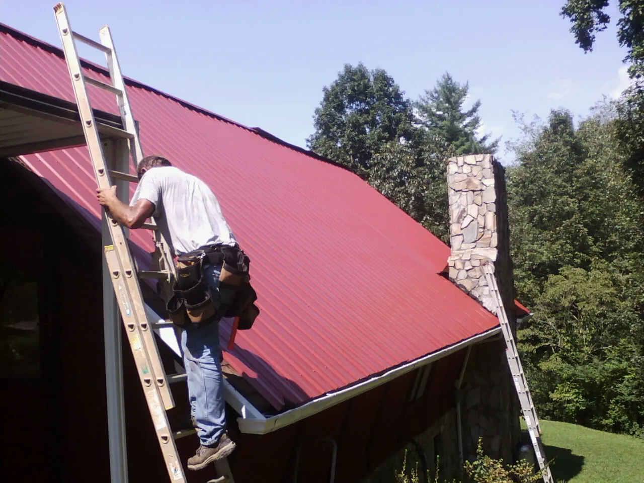 Roofer on a ladder beside a red metal roof and stone chimney — Soffit & Fascia Repair by Roofing Brothers in Larkspur